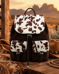 A brown and white cow-print backpack styled on a rustic wooden table with a cowboy hat, rope, and blanket, set against a desert landscape background at sunset.