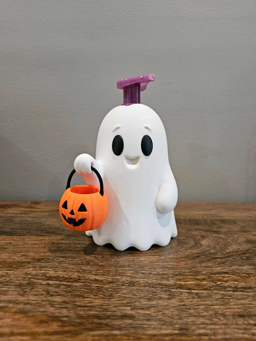 A white ghost-shaped pump dispenser with a purple top handle, holding a small orange jack-o'-lantern bucket, set on a wooden surface against a gray wall.