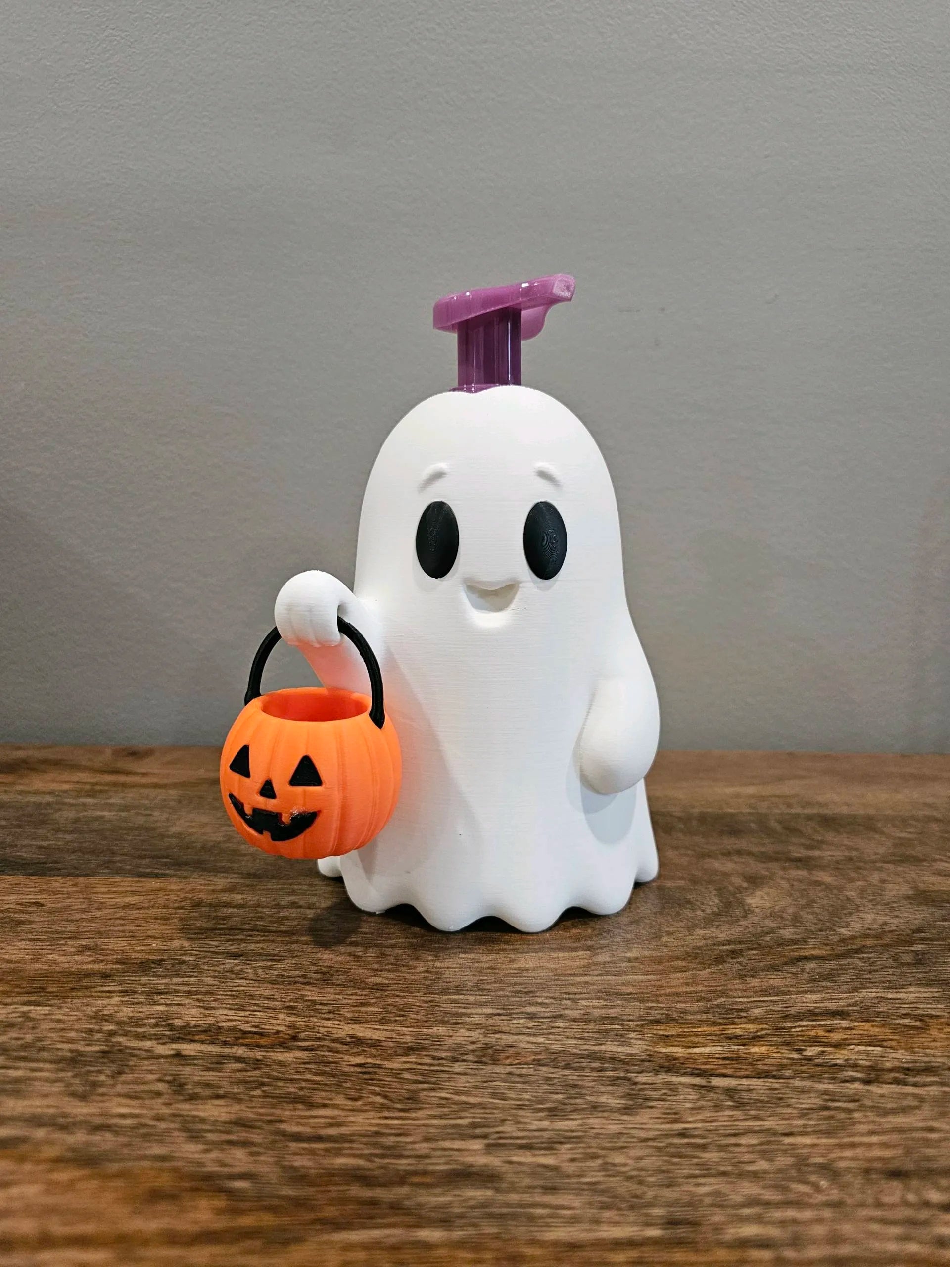 A white ghost-shaped pump dispenser with a purple top handle, holding a small orange jack-o'-lantern bucket, set on a wooden surface against a gray wall.