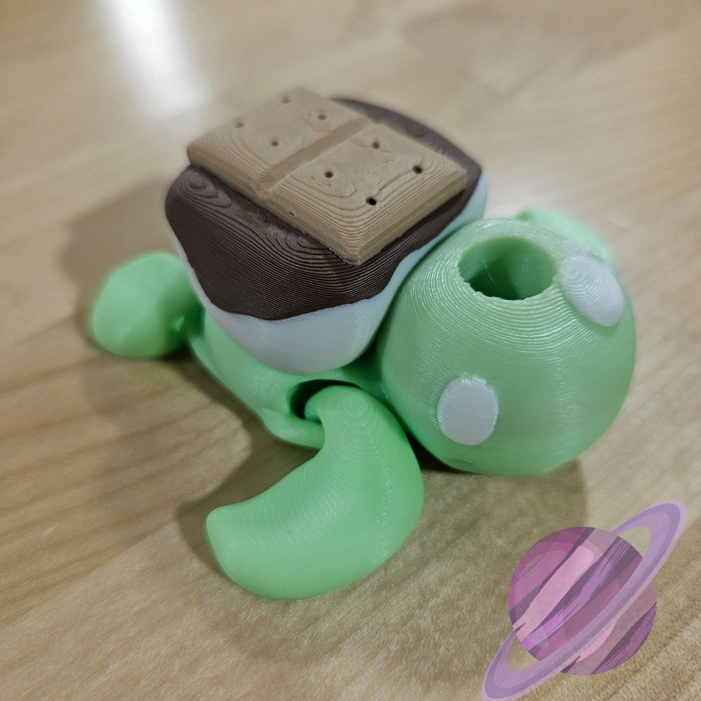 Toy figure of a green creature with a brown object on its back, resembling a s'more, on a wooden surface.