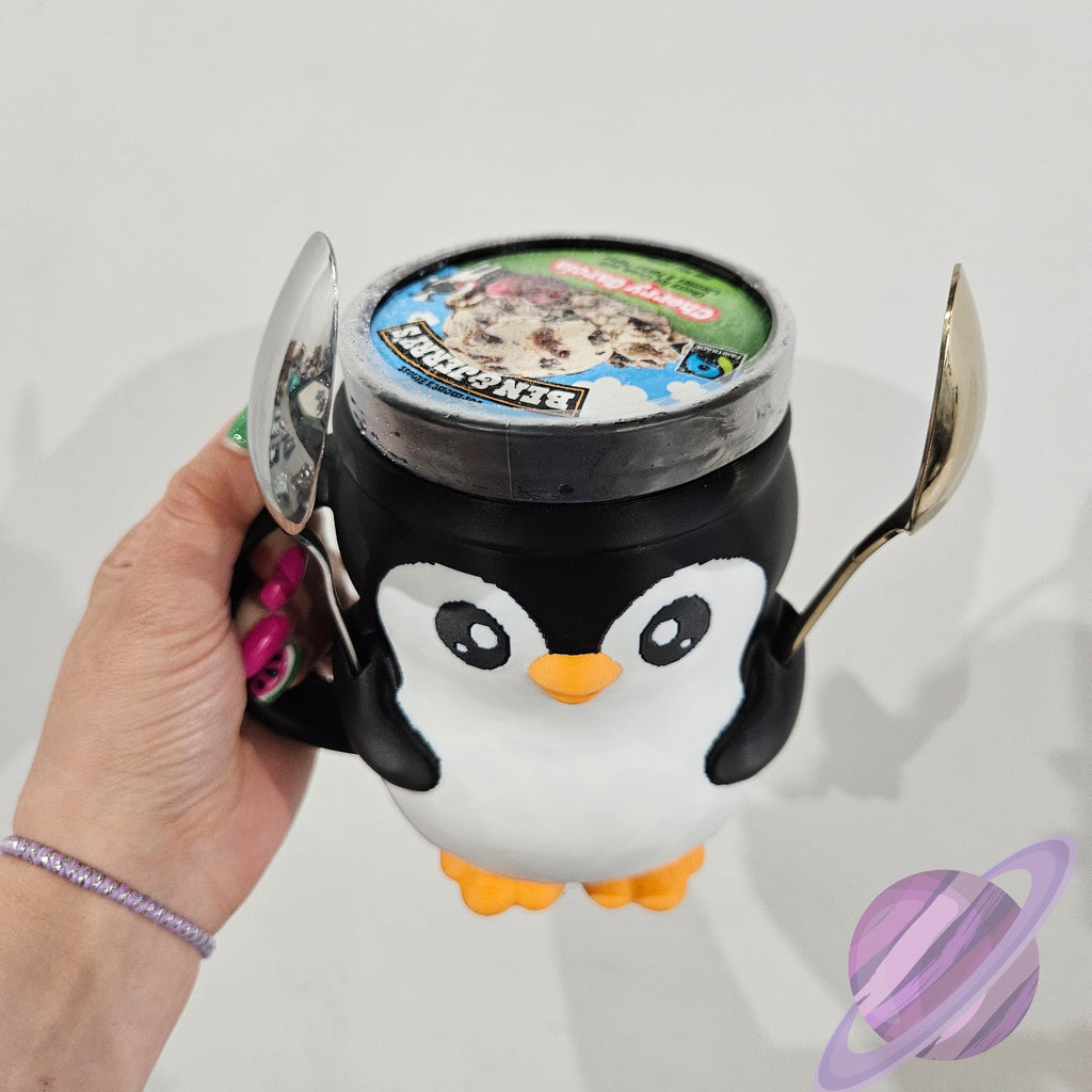 Penguin-shaped container with a lid and spoon, held by a hand against a light gray background.