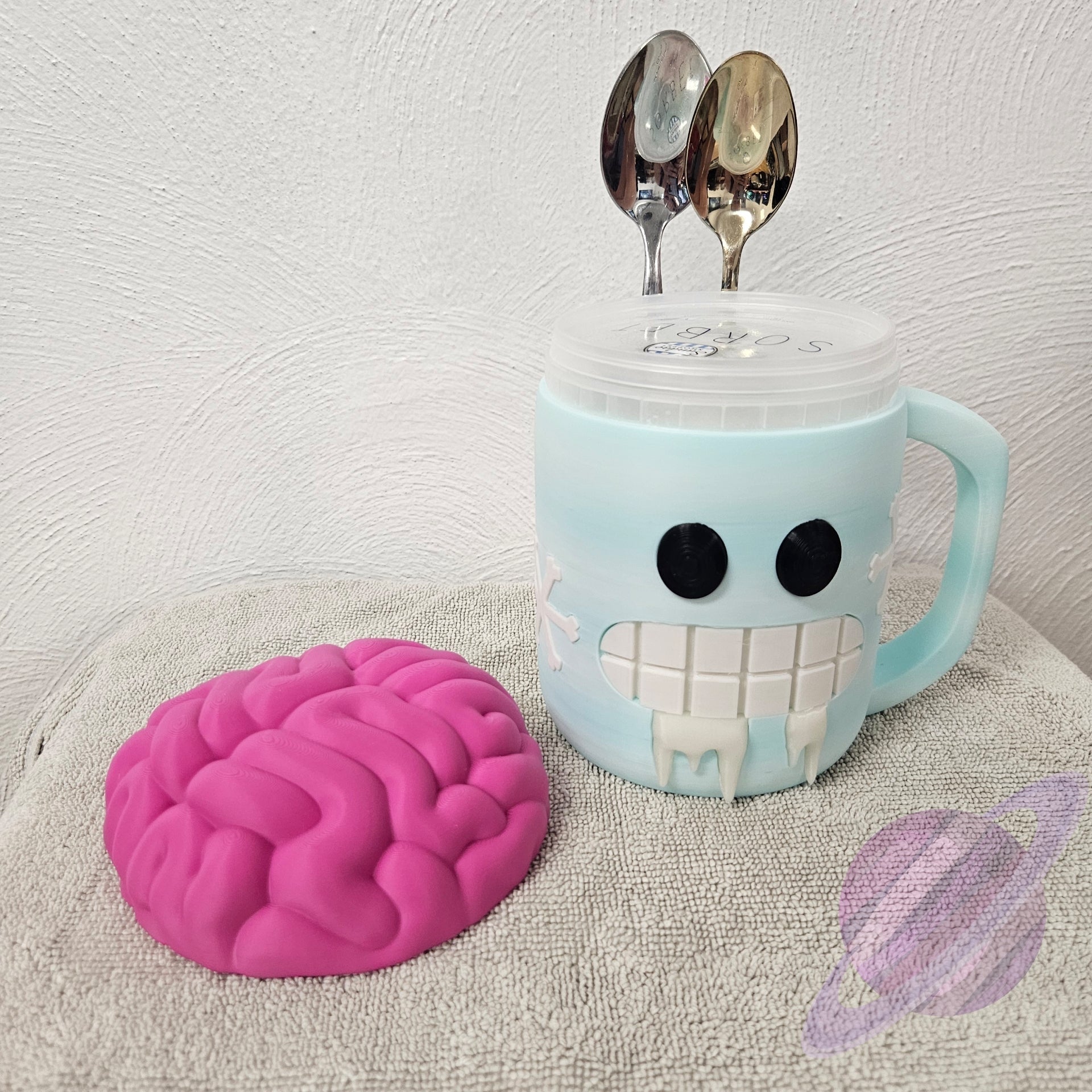 Blue mug with smiley face design, pink brain-shaped object, and two spoons on a textured surface.