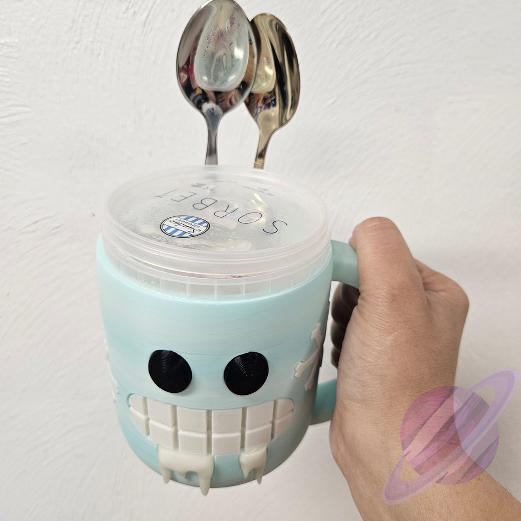 Light blue robot-shaped mug with a lid and two spoons on top, held by a hand against a white background.