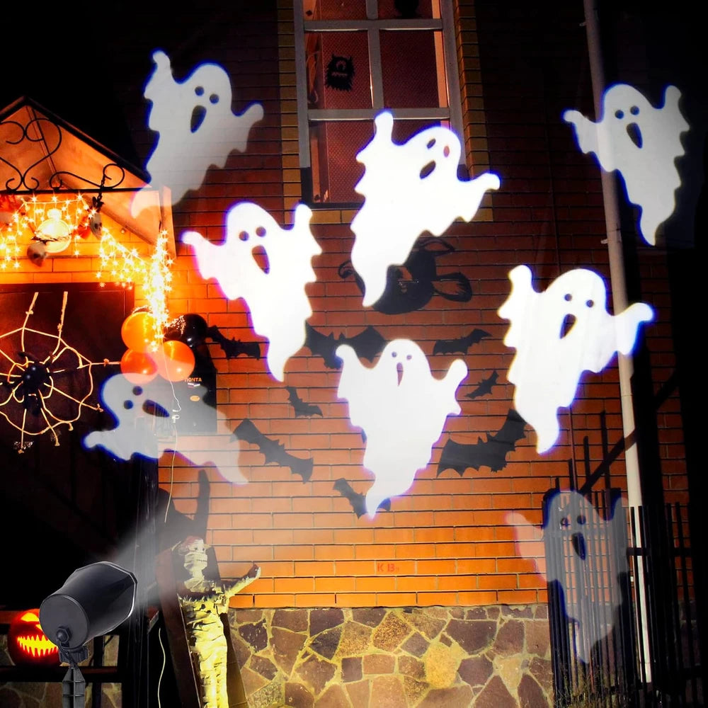 White ghost-shaped string lights projected onto a brick building facade at night, with orange window light and Halloween decorations visible in the background.