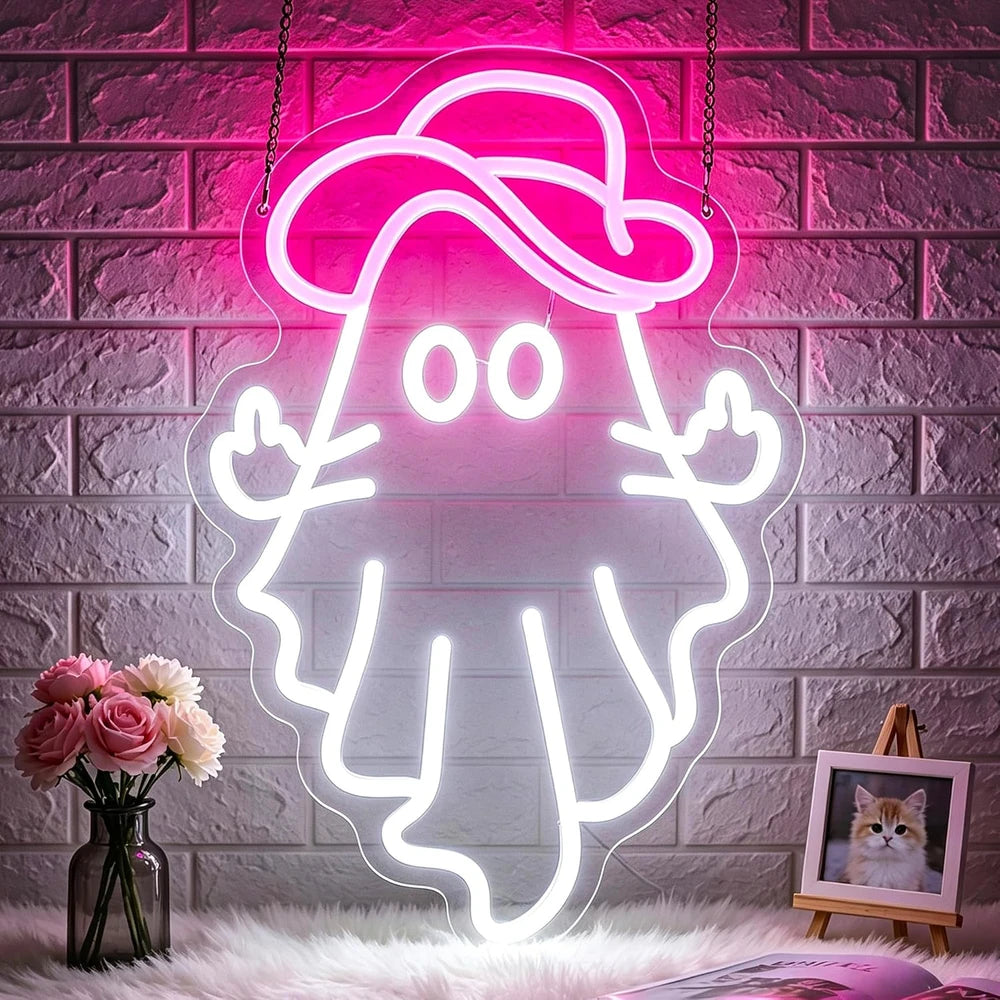 A glowing neon-style wall sign shaped like a cartoon ghost wearing a hat, mounted against a faux brick wall. The sign is illuminated in bright pink and white, with decorative flowers and a small framed picture on a surface below.
