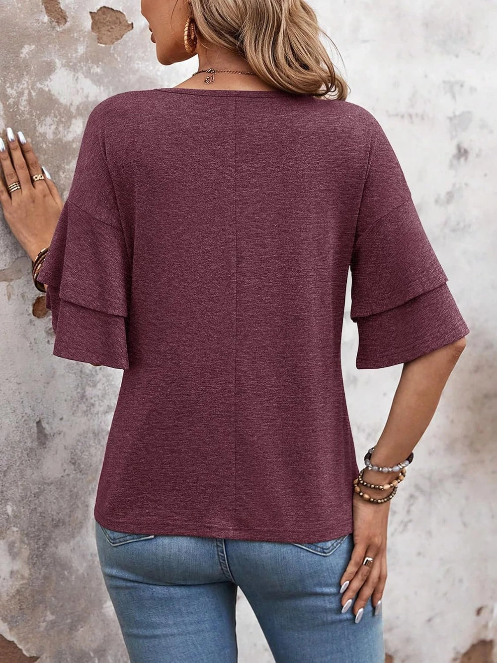 A woman shown from the back wearing a heathered burgundy top with layered flutter sleeves and blue jeans, standing against a textured concrete wall.