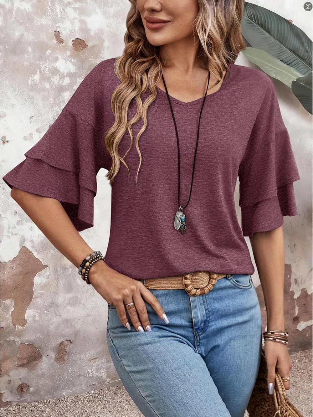 A woman wearing a heathered mauve-purple short-sleeve top with layered ruffle cuffs, paired with light blue jeans and a long pendant necklace, posing against a textured plaster wall.