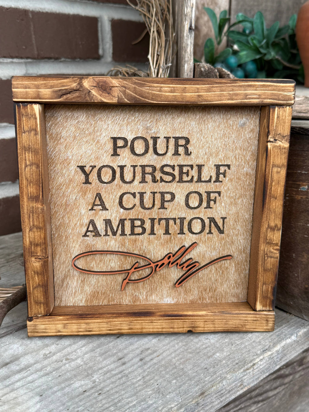 A rustic wooden framed sign with the phrase 'POUR YOURSELF A CUP OF AMBITION' and a cursive signature reading 'Dolly', displayed on a wooden surface with a brick wall and plants in the background.
