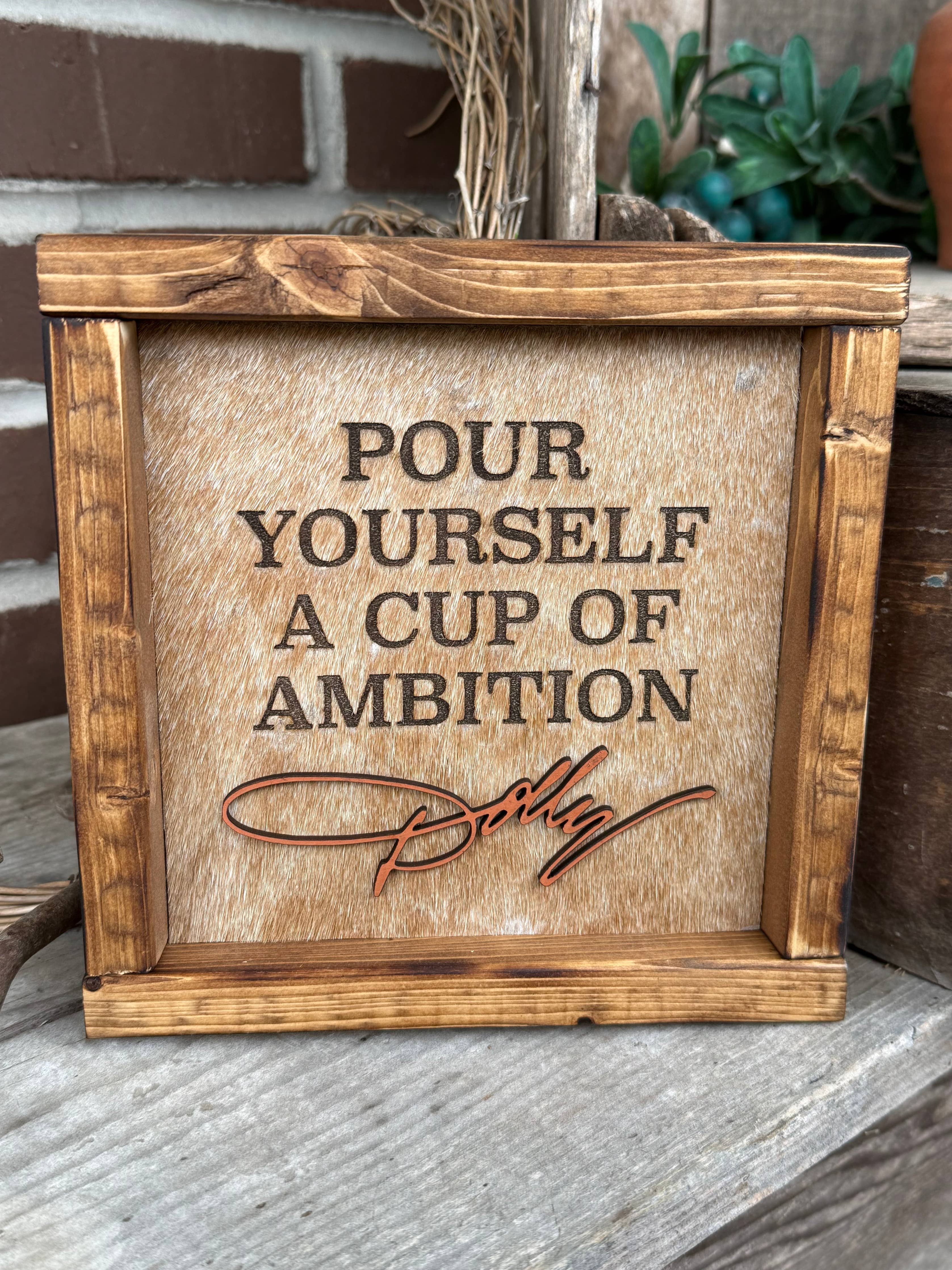 A rustic wooden framed sign with the phrase 'POUR YOURSELF A CUP OF AMBITION' and a cursive signature reading 'Dolly', displayed on a wooden surface with a brick wall and plants in the background.