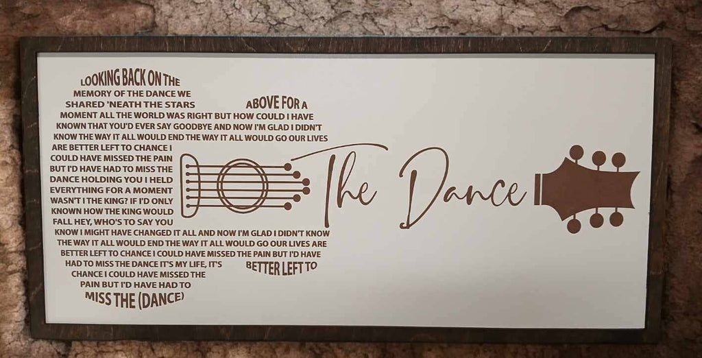A rectangular white wall plaque with printed lyrics and guitar graphics, mounted on a dark wood background. The central text reads 'The Dance' in large script, with song lyrics and guitar chord diagrams printed around it.