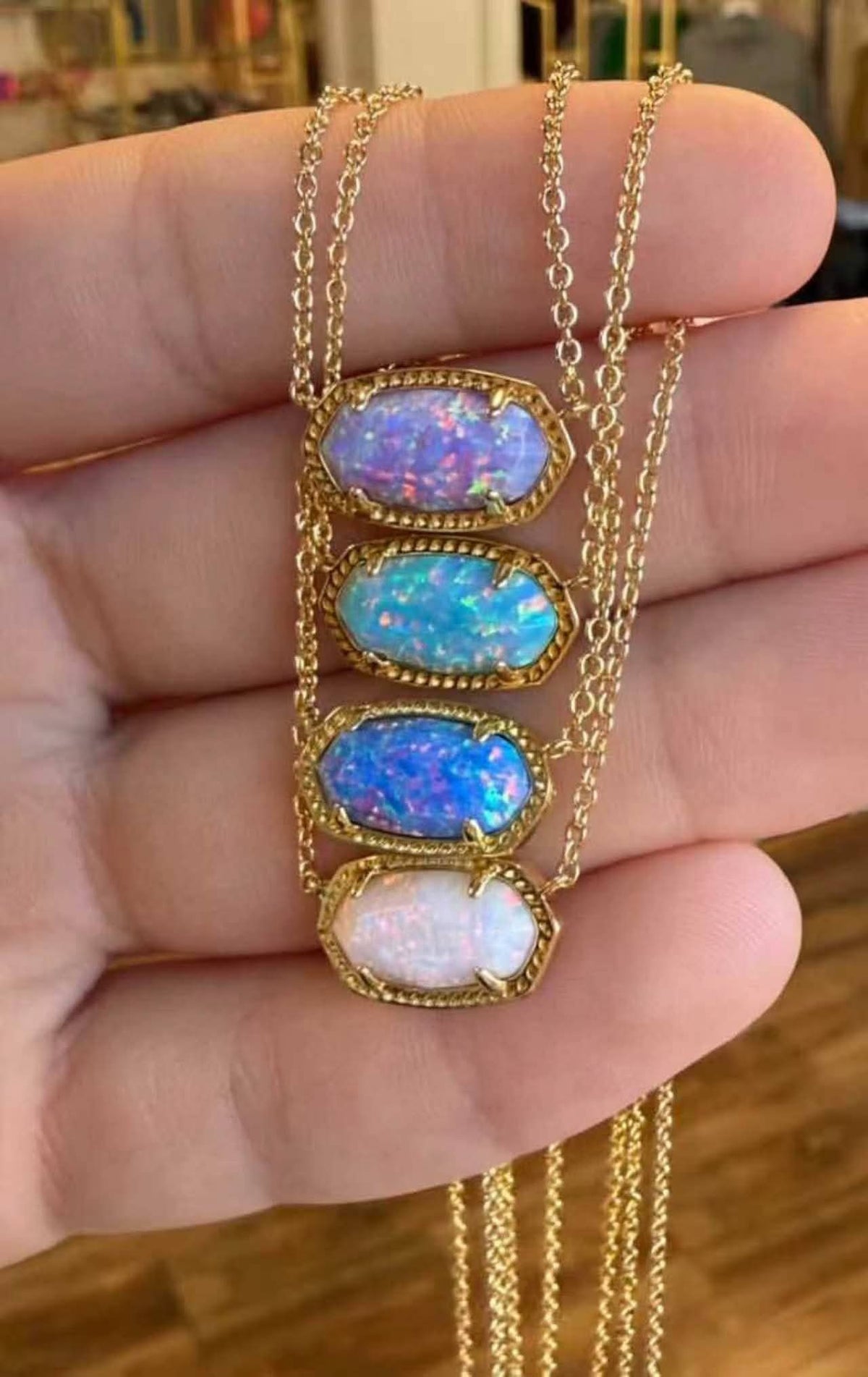 A gold chain necklace with four stacked oval opal-like pendants in different iridescent colors, held in a person's hand against a blurred indoor background.