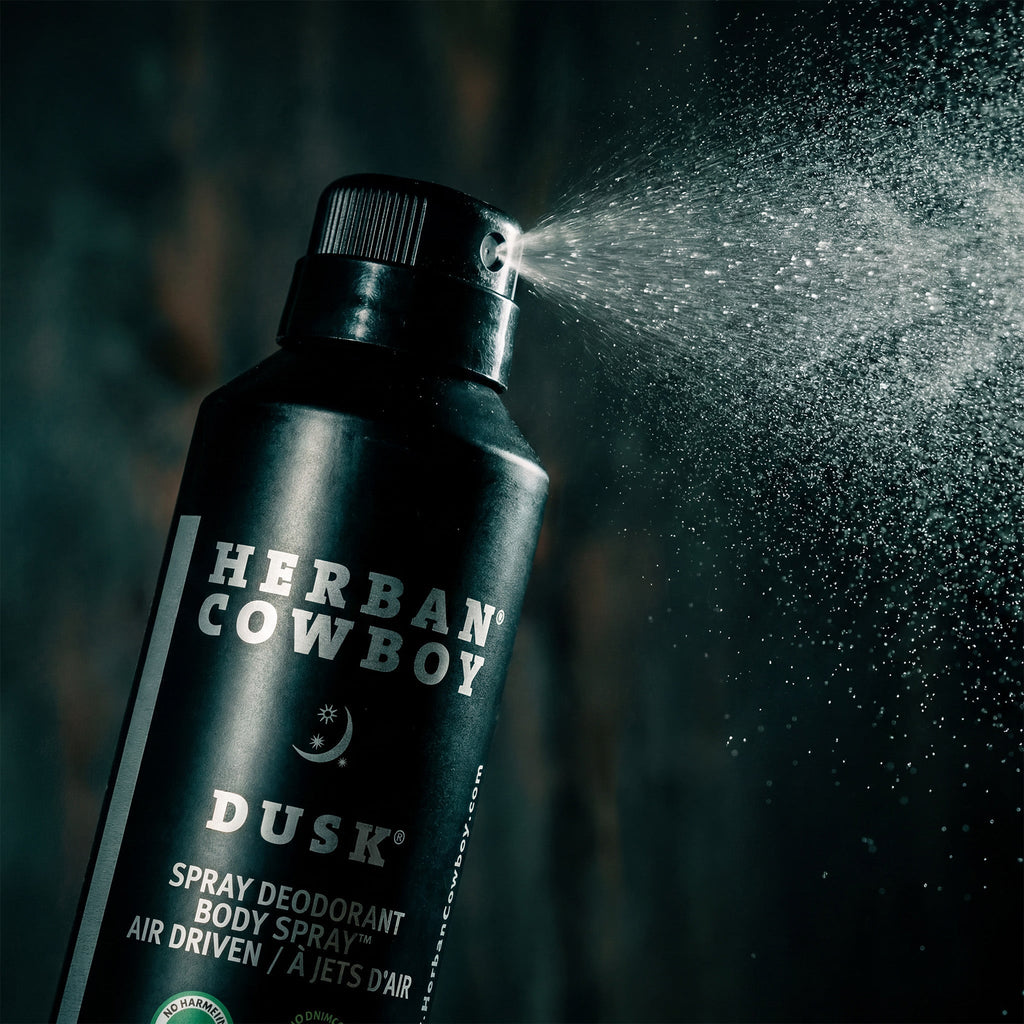 Herban Cowboy Dusk spray deodorant being sprayed against a dark background