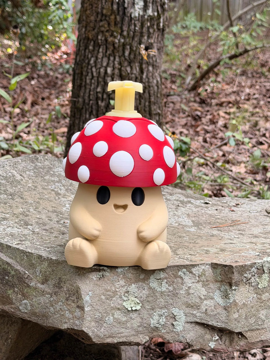 A cute mushroom-shaped soap dispenser or bottle with a red cap, white polka dots, and a beige body, sitting on a stone in an outdoor forest setting.
