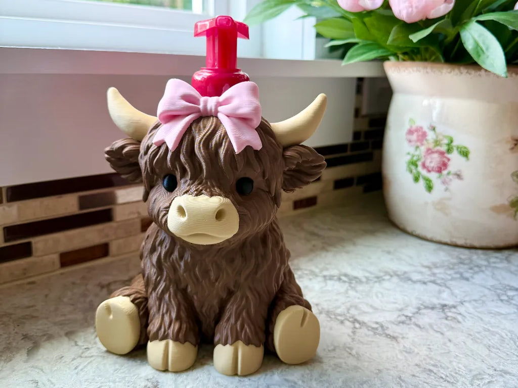 A brown Highland cow-shaped soap dispenser with a pink bow and pink pump, sitting on a kitchen counter next to a potted plant.