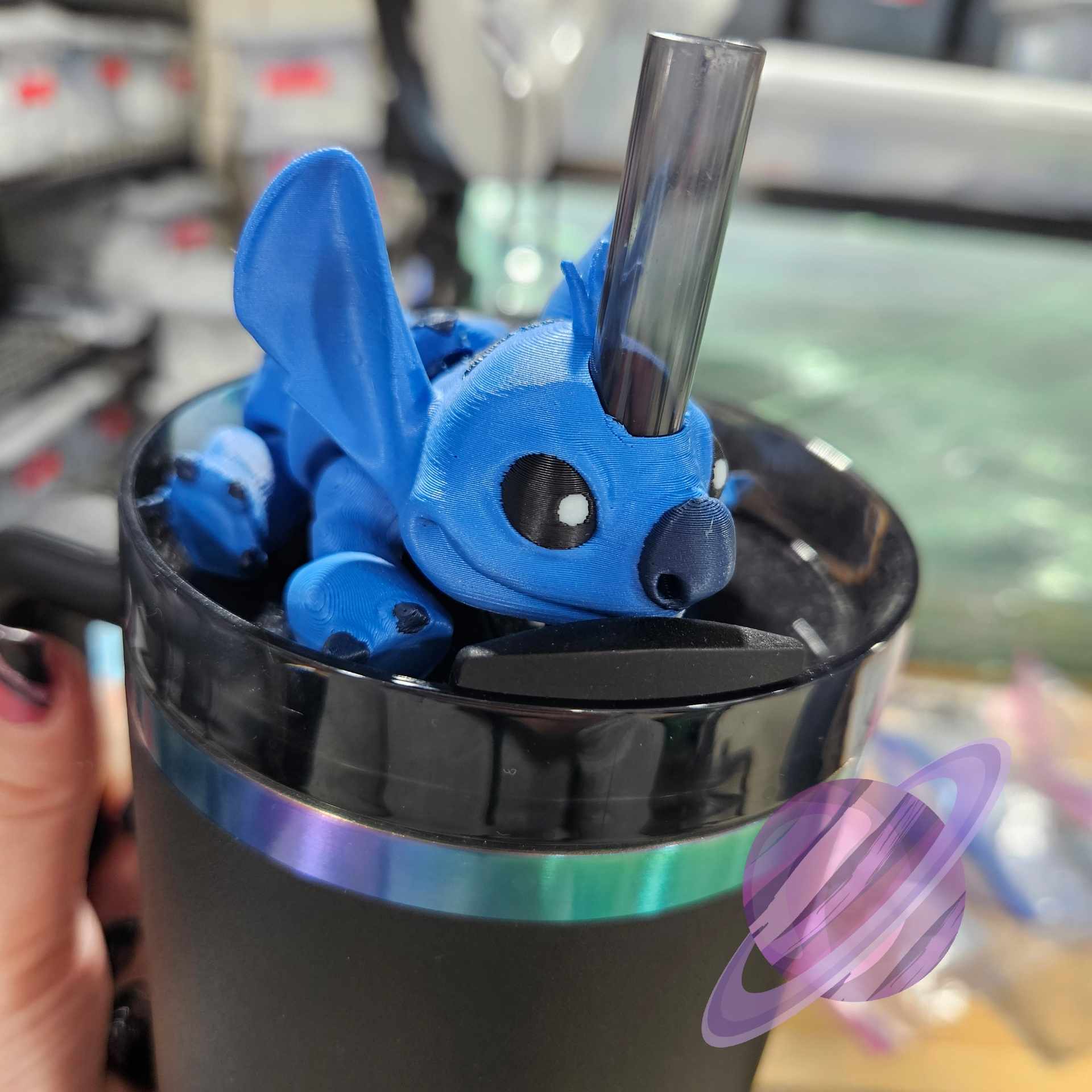 Blue plush toy with a straw in a black cup against a blurred indoor background