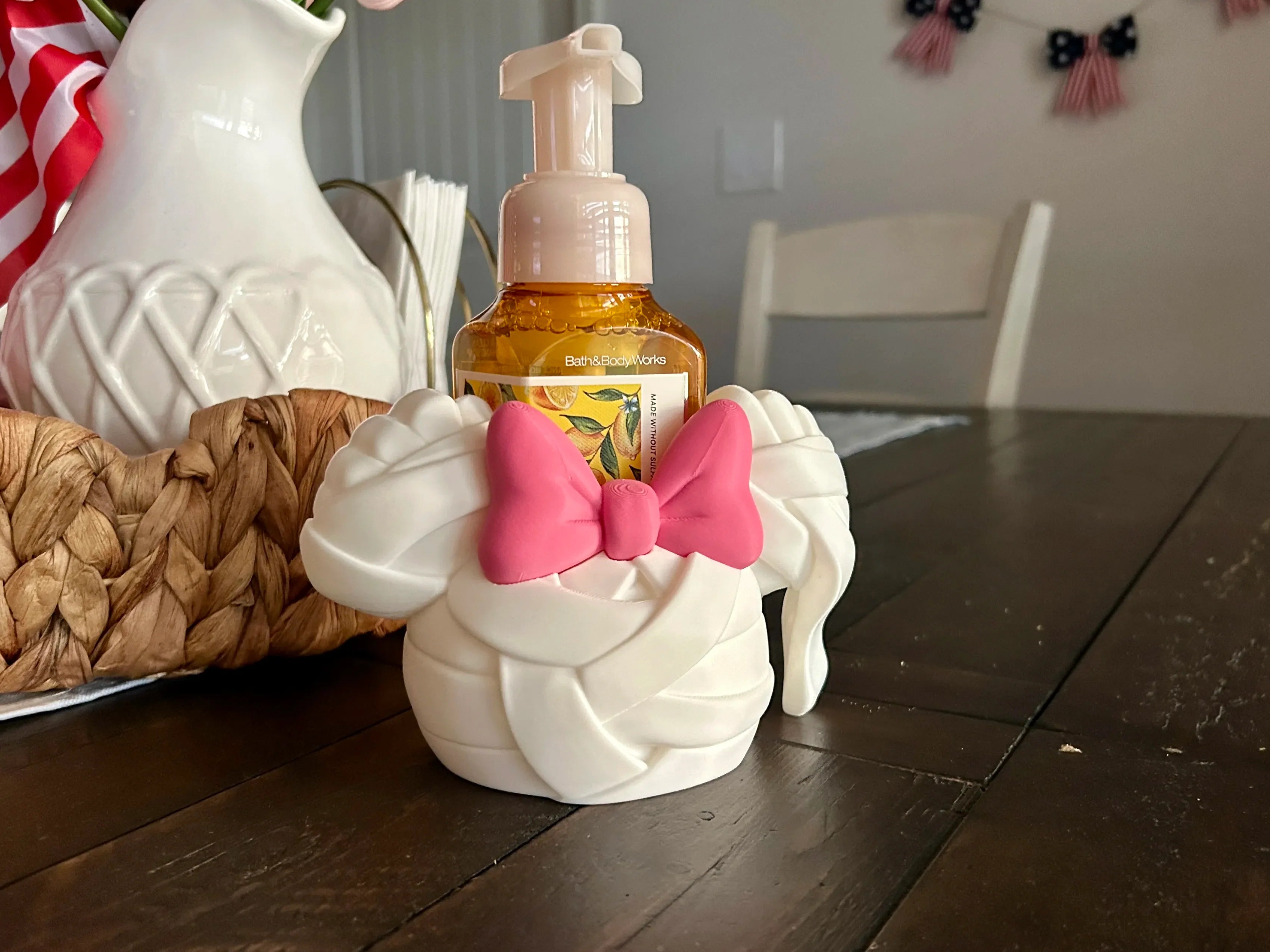 A white decorative foam dispenser shaped like a braided hair bun with a pink bow, holding a bottle of handcrafted soap on a dark wooden table.