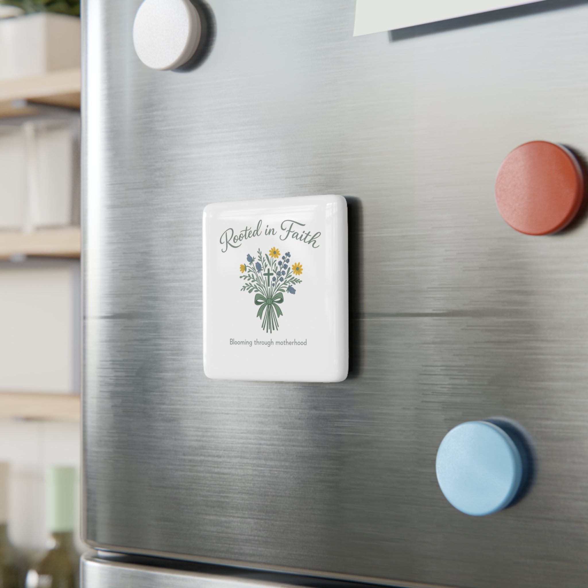 A square white refrigerator magnet with a floral bouquet illustration and the words 'Rested in Faith' and 'Blooming through adversity' is shown attached to a brushed stainless steel fridge door, alongside other colorful magnets.