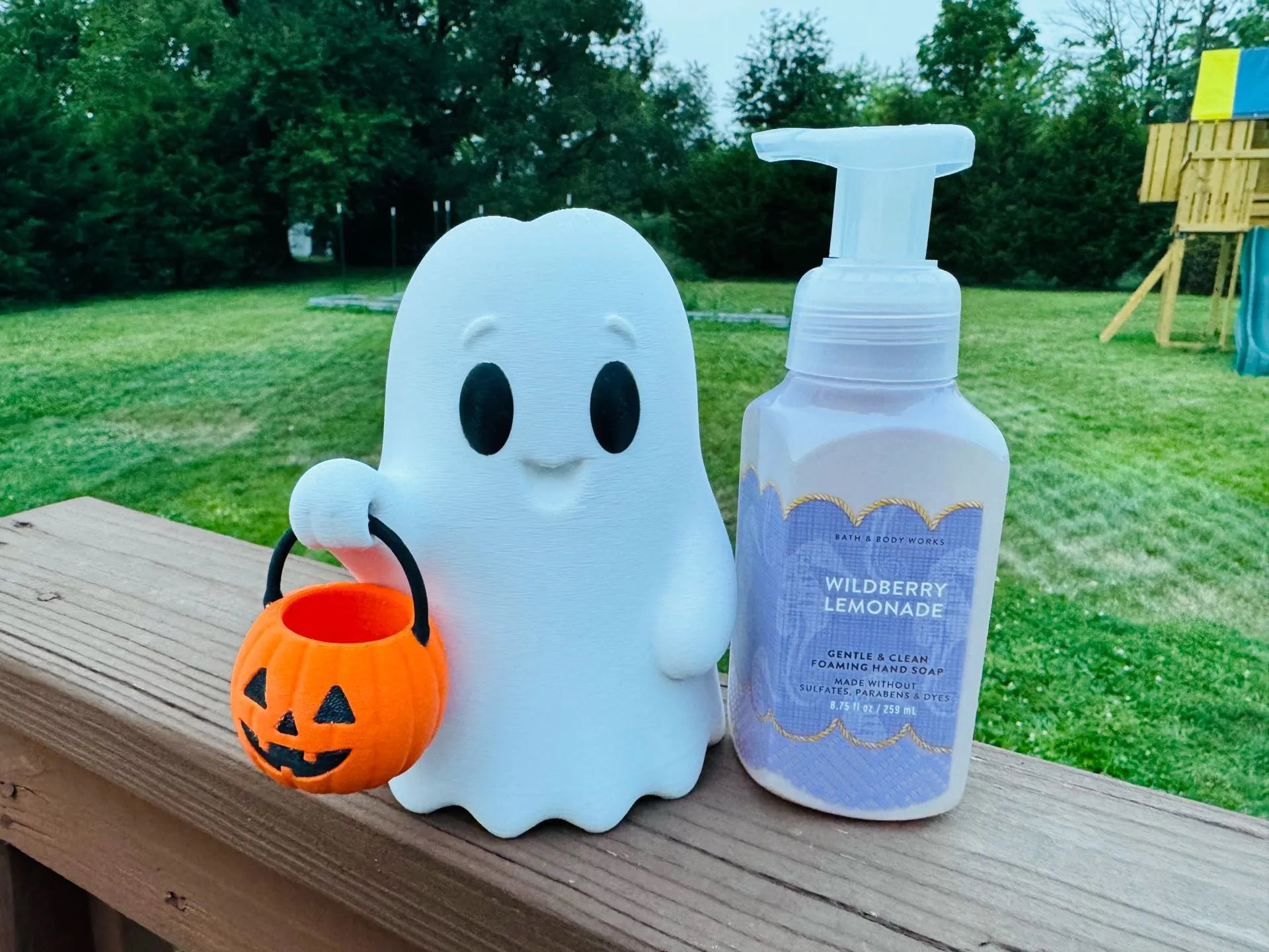 A white ghost-shaped container holding an orange pumpkin bucket sits next to a pump bottle labeled Wildberry Lemonade on a wooden deck with a grassy backyard in the background.