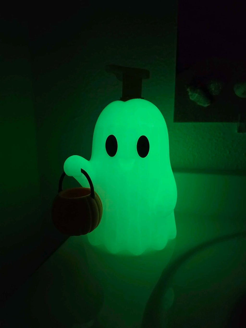 A glowing green ghost-shaped night light with black eyes and a small pumpkin bucket accessory, shown in a dark room.