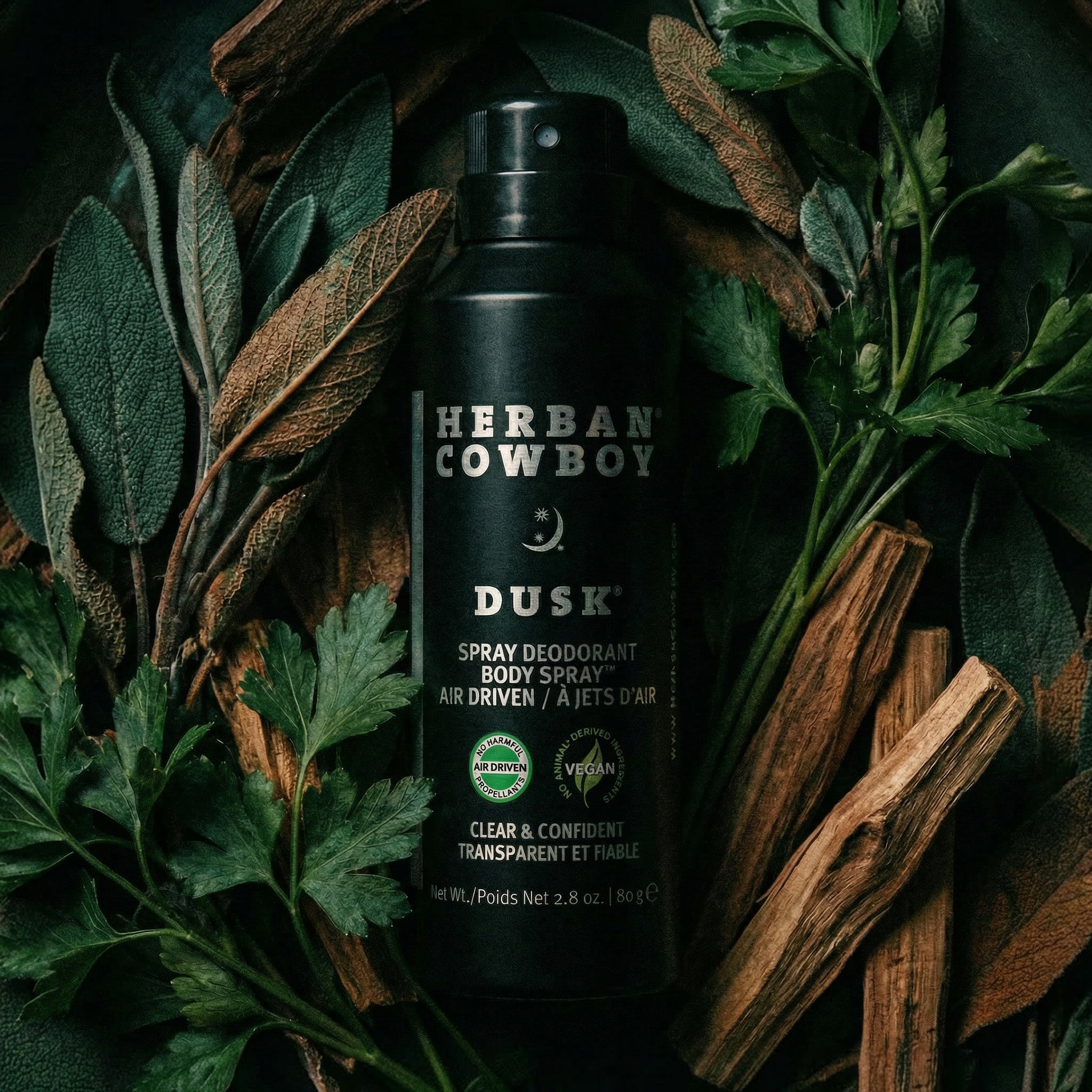 Herban Cowboy Dusk spray deodorant surrounded by leaves and wood on a dark background