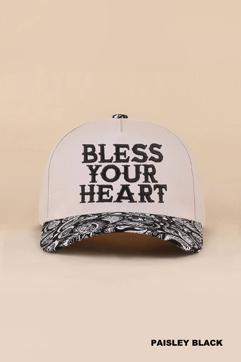 A white baseball cap with 'BLESS YOUR HEART' printed in black on the front, featuring a paisley-patterned black and white brim, set against a solid beige background.