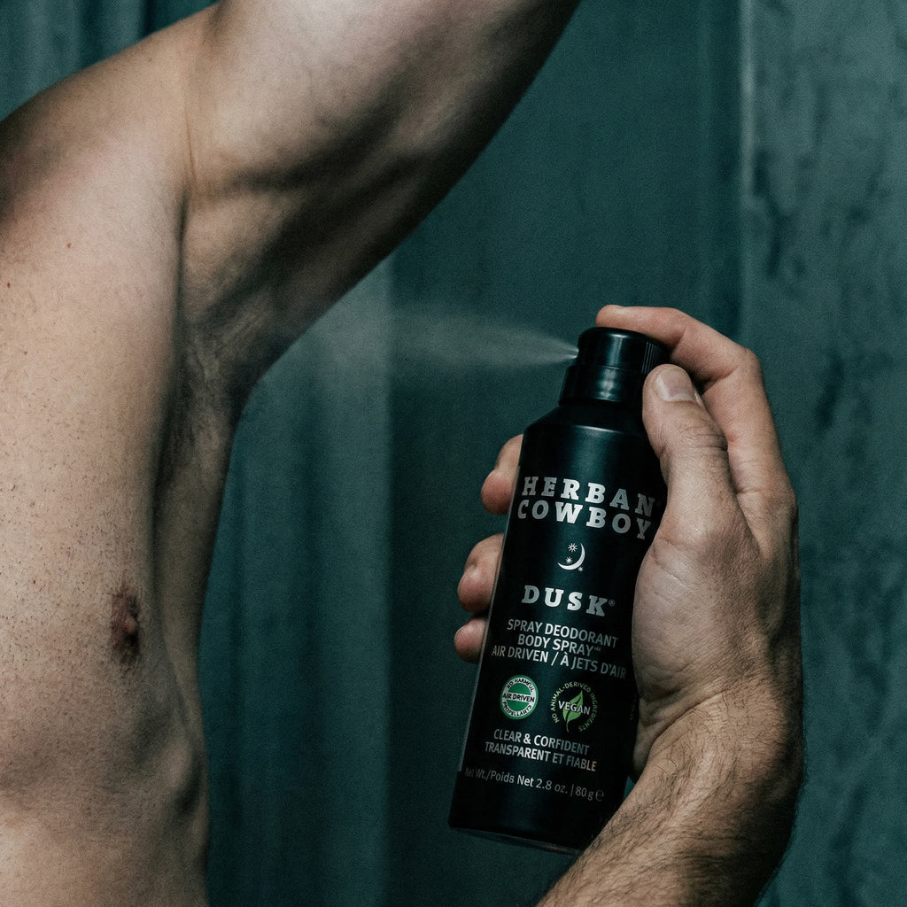 Person holding a bottle of 'Herbal Cowboy' Dusk spray deodorant against a dark background