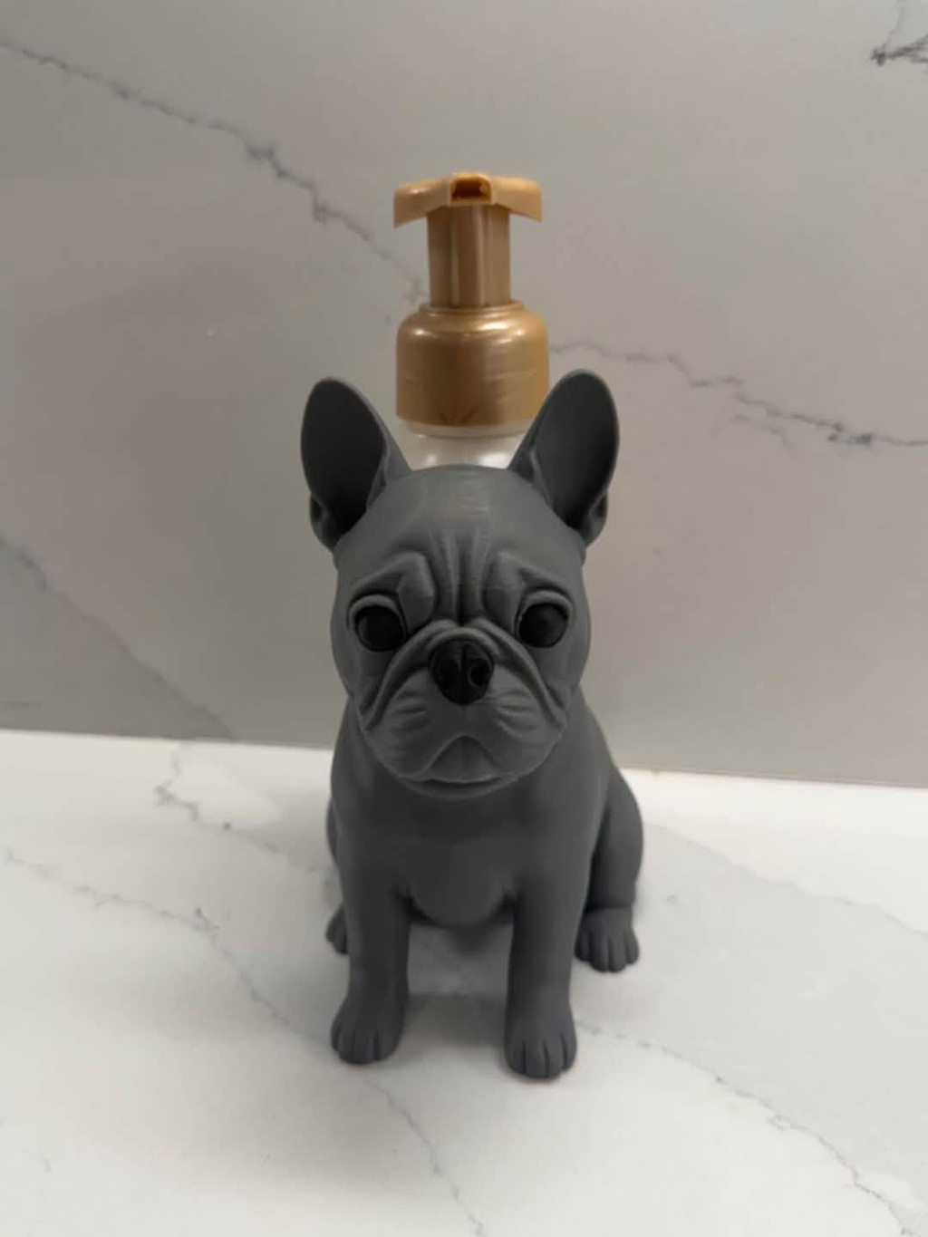 A gray French Bulldog-shaped pump bottle with a gold-colored pump dispenser, set against a white marble-patterned background.