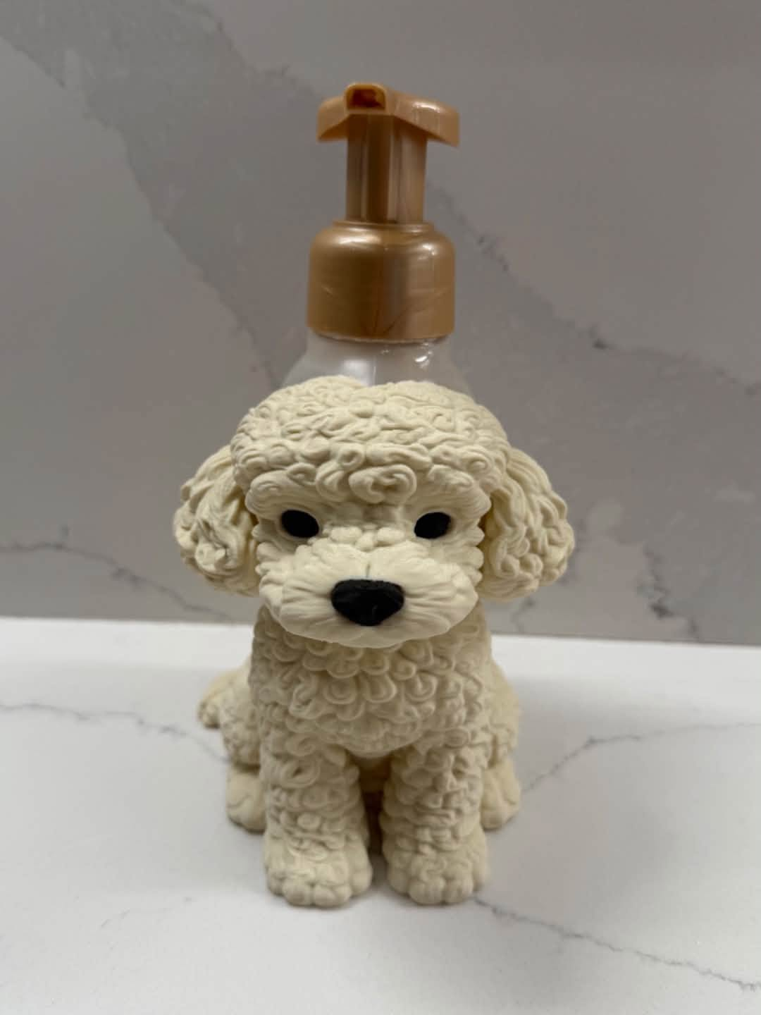 A small cream-colored poodle figurine with a glossy black nose and eyes, mounted on a tan pump dispenser, set against a gray marble-patterned background.