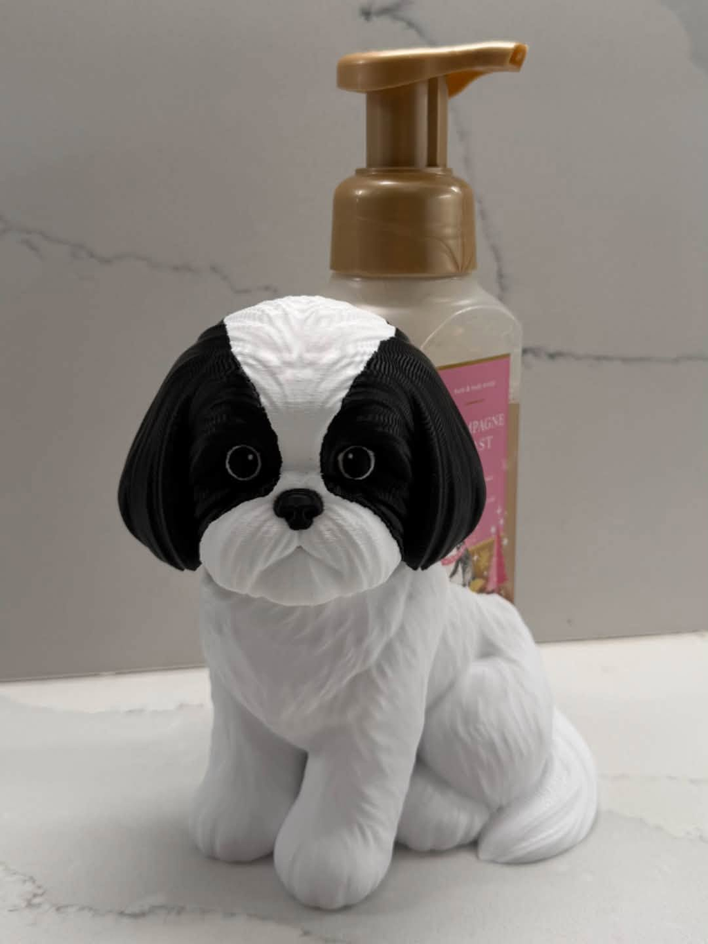 A small black-and-white dog figurine with a glossy finish, styled as a dog-shaped bottle holder in front of a pump bottle on a light marble surface.