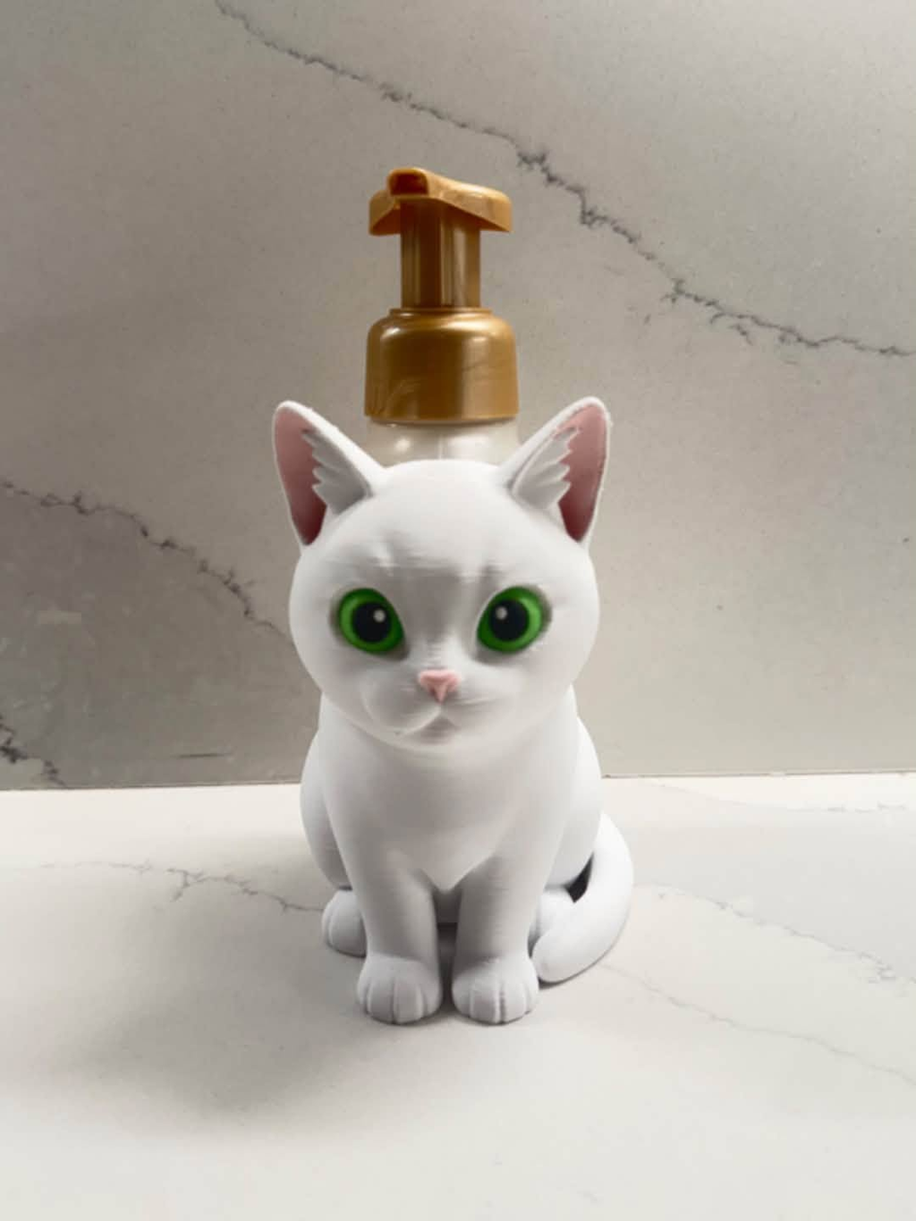 A white cat-shaped pump bottle with large green eyes and a metallic gold pump dispenser, set against a light gray marble-patterned background.