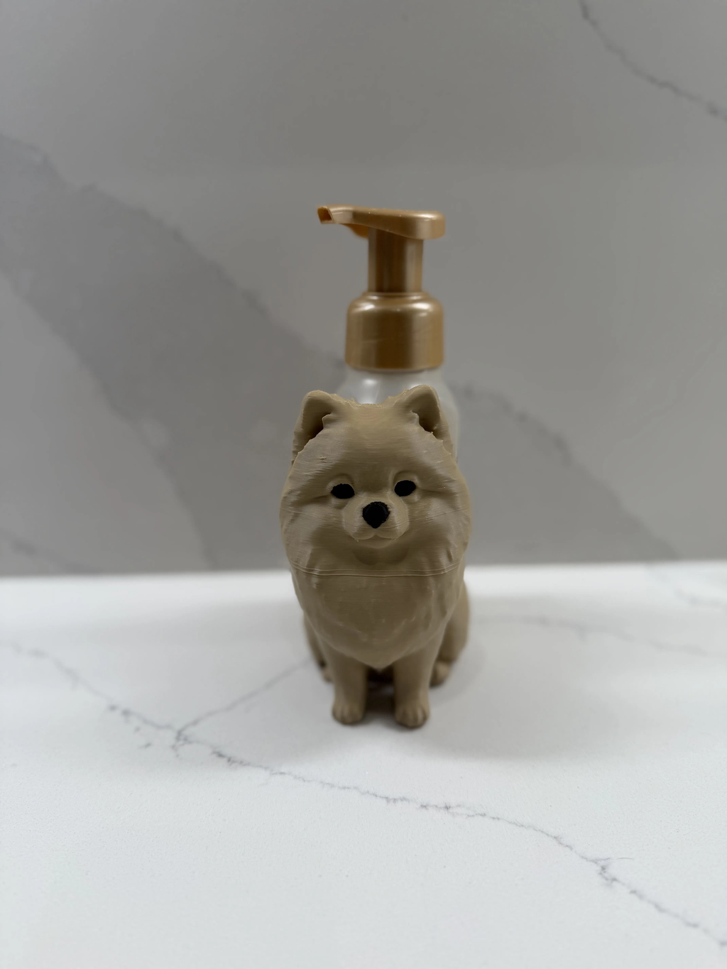 A small dog-shaped pump bottle with a beige body and a metallic gold pump dispenser, standing on a white marble-like surface against a gray marble-patterned background.