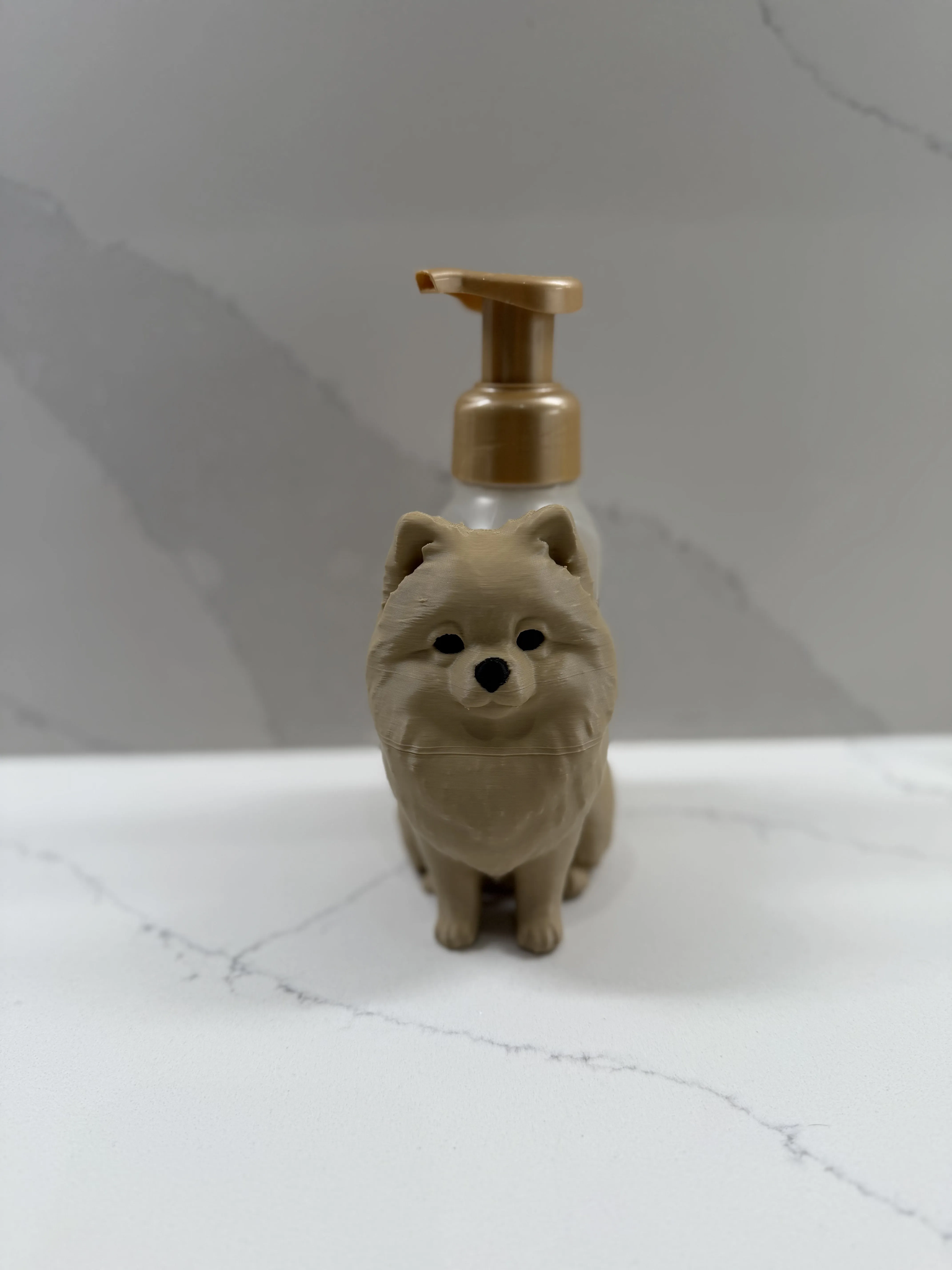 A small dog-shaped pump bottle with a beige body and a metallic gold pump dispenser, standing on a white marble-like surface against a gray marble-patterned background.