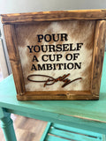 A rustic wooden sign with a cowhide panel, featuring the engraved text 'POUR YOURSELF A CUP OF AMBITION' and a cursive signature that reads 'Dolly'. The sign is displayed on a distressed teal painted table.
