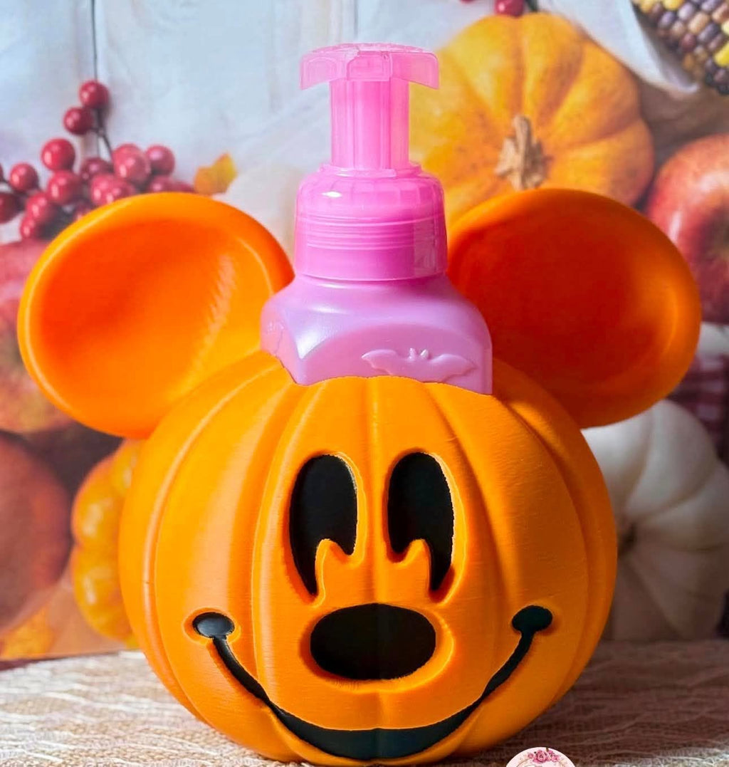 An orange Mickey Mouse-shaped Halloween jack-o'-lantern pump bottle with a pink pump dispenser, set against a blurred autumnal background with pumpkins and red berries.