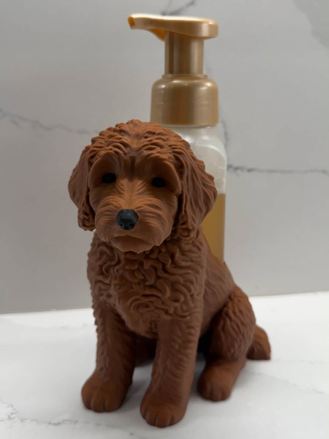 A brown dog-shaped pump dispenser or holder positioned in front of a gold-and-clear pump bottle on a light marble-like surface and wall.