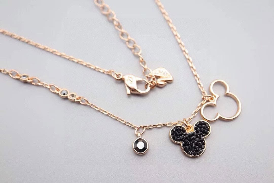 A rose gold necklace featuring a Mickey Mouse head charm covered in black crystals, a Mickey Mouse ear charm, a small round black gem, and a heart-shaped charm, displayed on a light gray background.