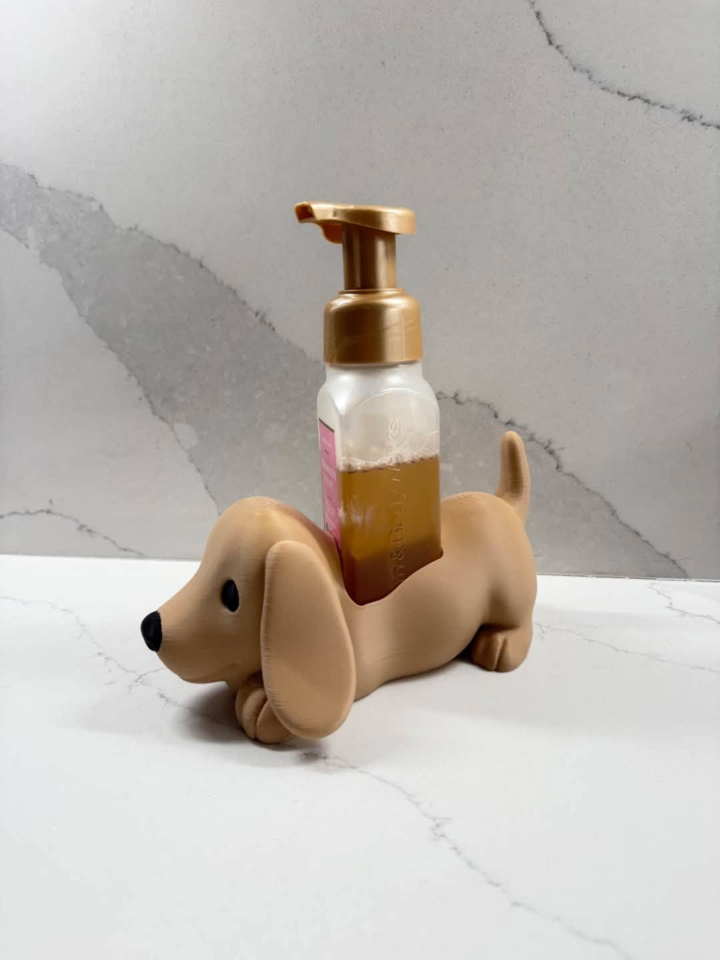 A small tan dachshund-shaped bottle holder or stand holding a frosted pump bottle filled with amber liquid, set on a white marble-like surface against a gray marble-patterned background.