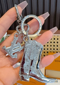 A hand with long nude acrylic nails holds a silver-tone metal keychain featuring a cowboy boot charm and a monogram keyring.