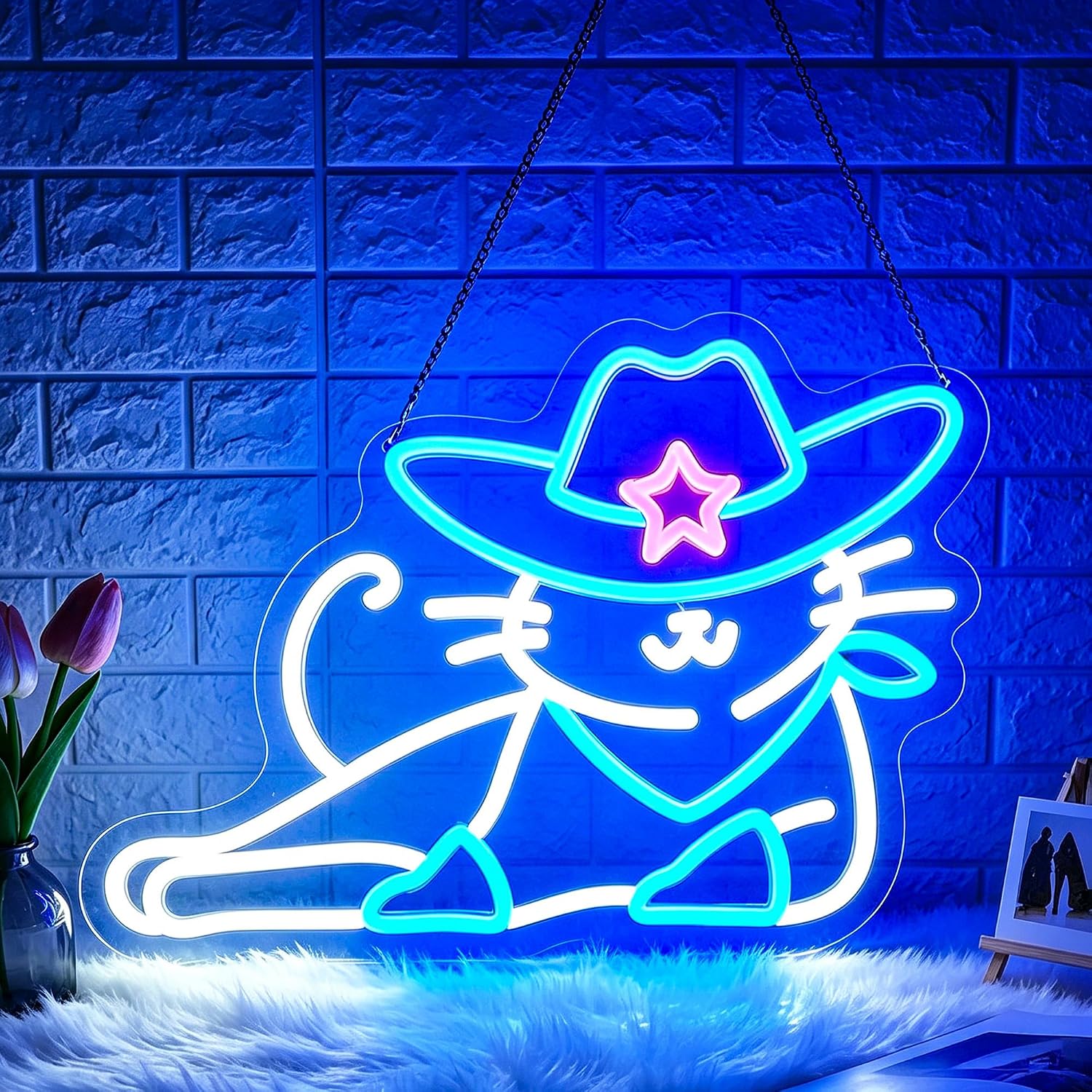 A neon-style wall sign shaped like a cat wearing a cowboy hat, glowing in bright blue, white, and pink tones against a blue brick wall. The sign is hanging by chains, with decorative faux fur at the bottom and small props including tulips and a small picture frame nearby.
