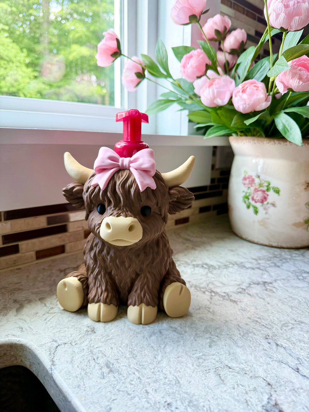 A brown highland cow soap dispenser with a pink bow and red pump, sitting on a kitchen counter next to a vase of pink flowers.