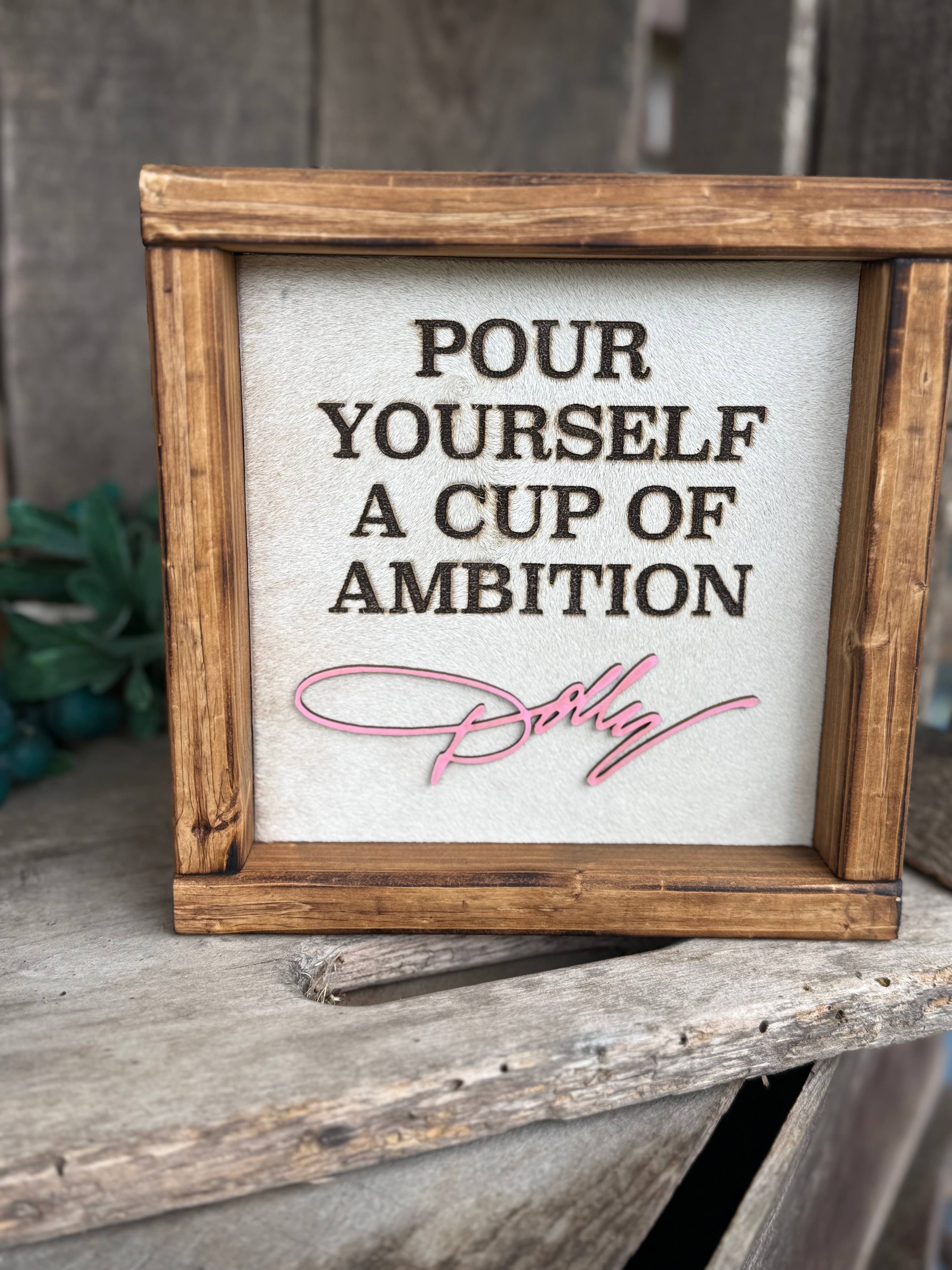 A square framed decorative sign with the phrase “POUR YOURSELF A CUP OF AMBITION” in dark lettering on a light background, with a pink script signature reading “Betsy” near the bottom. The sign is set in a rustic wooden frame and displayed on a weathered wooden surface.