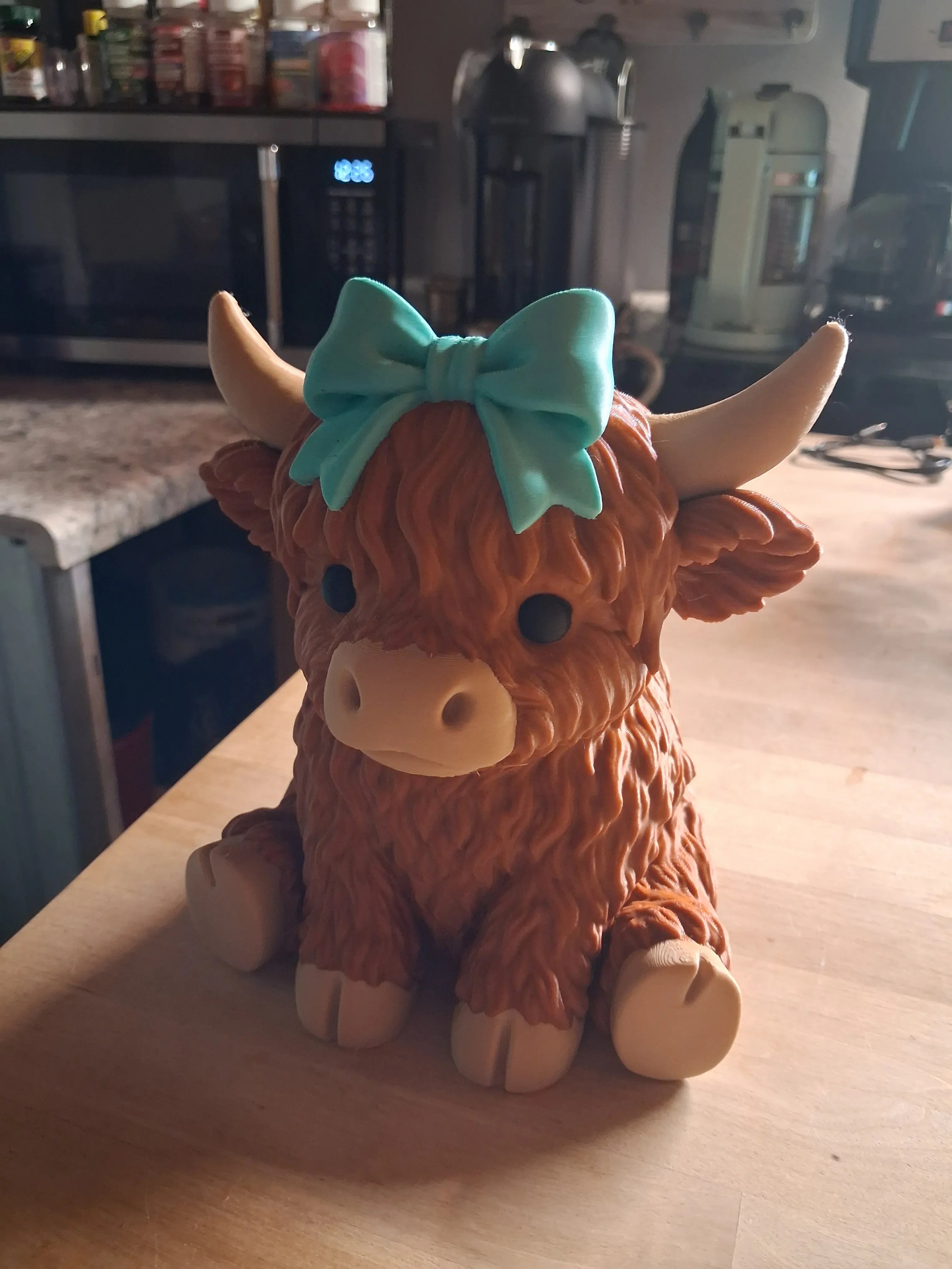 A Highland cow figurine with shaggy brown fur, cream-colored horns and hooves, and a bright turquoise bow on its head, sitting on a wooden table in a kitchen setting.