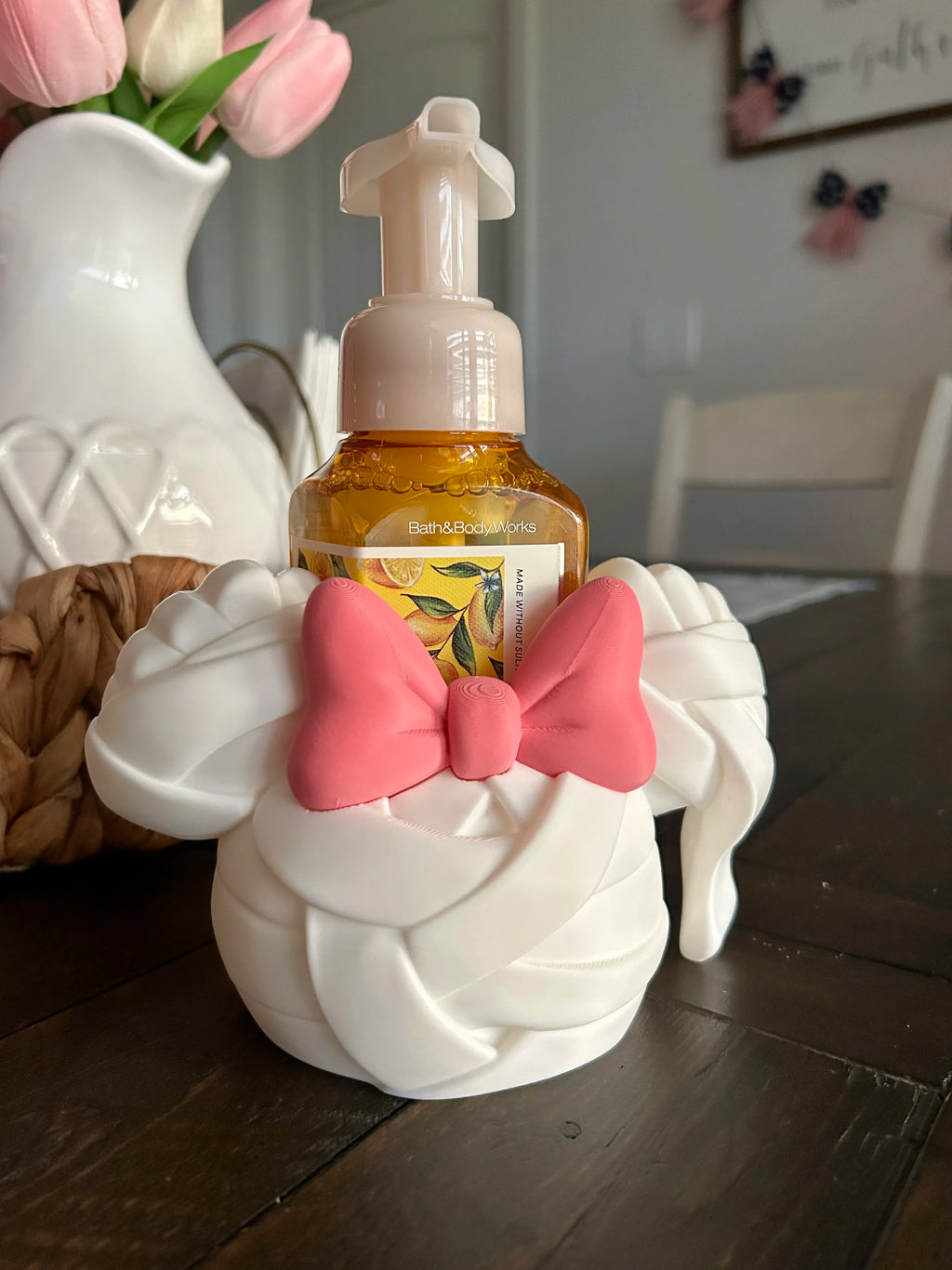 A white decorative soap dispenser shaped like a twisted towel or ribbon, with a pink bow on the front, holding a small amber bottle with a pump. The dispenser sits on a dark wooden table in a home setting, with a white pitcher of pink tulips blurred in the background.