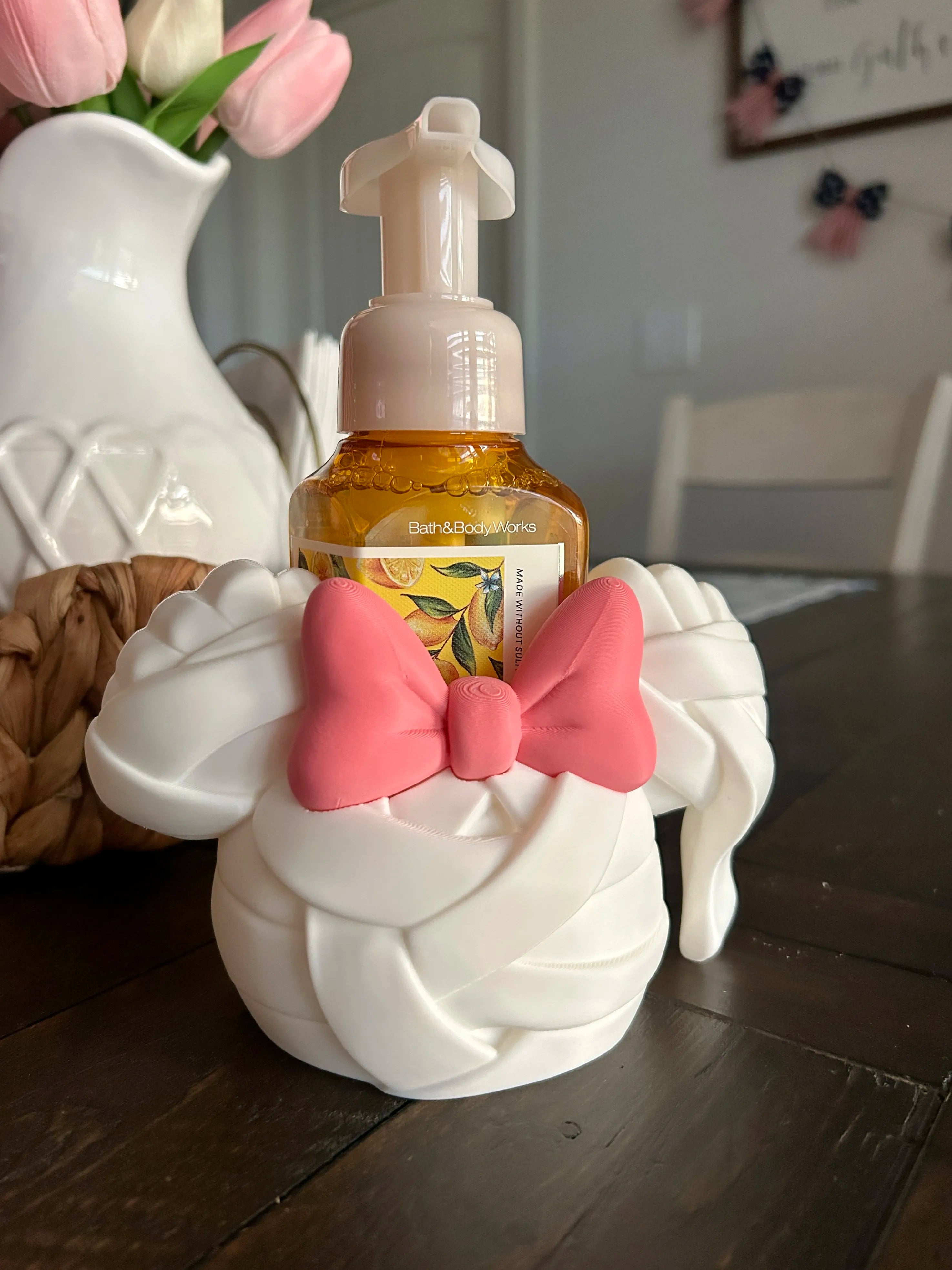 A white decorative soap dispenser shaped like a twisted towel or ribbon, with a pink bow on the front, holding a small amber bottle with a pump. The dispenser sits on a dark wooden table in a home setting, with a white pitcher of pink tulips blurred in the background.