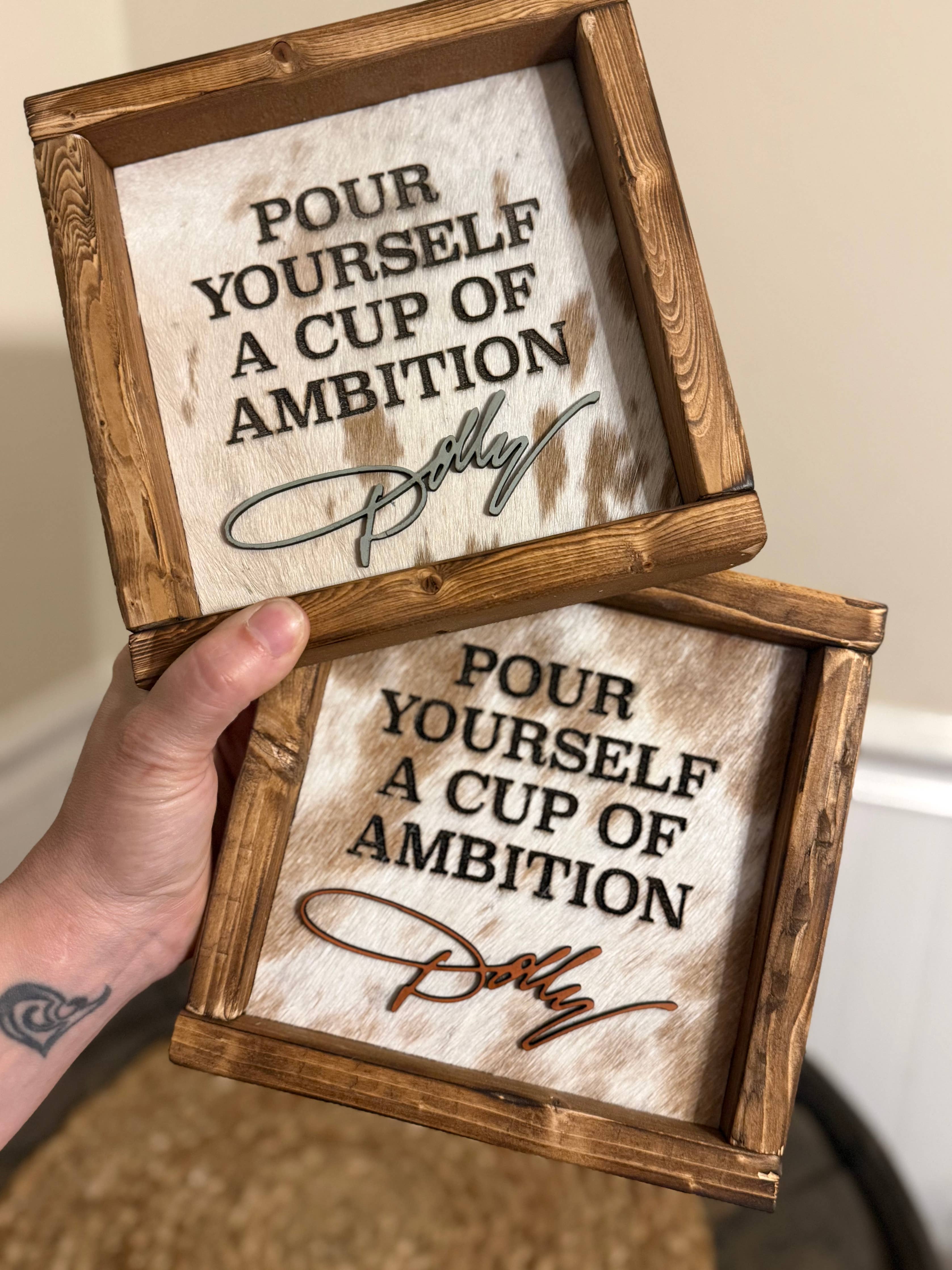 Two square wooden framed signs with the phrase 'Pour Yourself a Cup of Ambition' and a cursive signature, held by a hand against an indoor background.