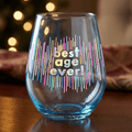Clear glass with 'best age ever!' text on a wooden surface with blurred lights in the background