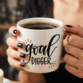 Person holding a mug with 'goal digger' text in a blurred cafe setting
