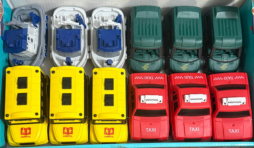 A boxed set of small toy vehicles arranged in a tray inside an open cardboard box, photographed from above on a chair with a wooden floor visible. The set includes yellow, blue, red, and green toy cars and trucks.