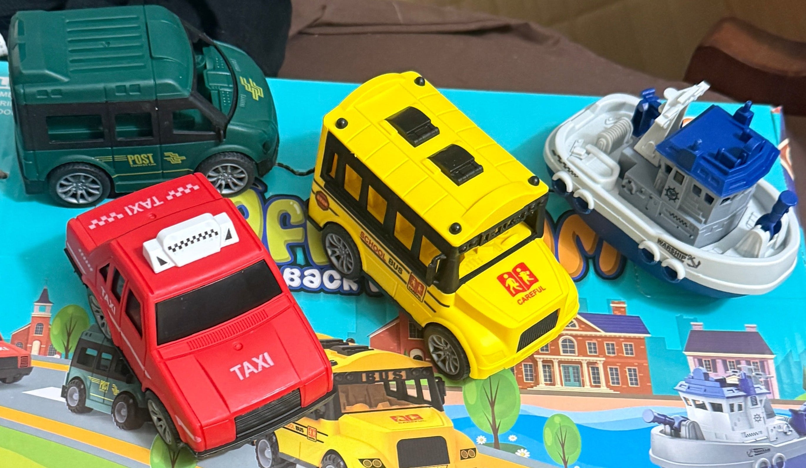 A set of colorful toy vehicles, including a red taxi, yellow school bus, green truck, and a white boat, displayed on a printed toy road mat on a brown chair cushion.