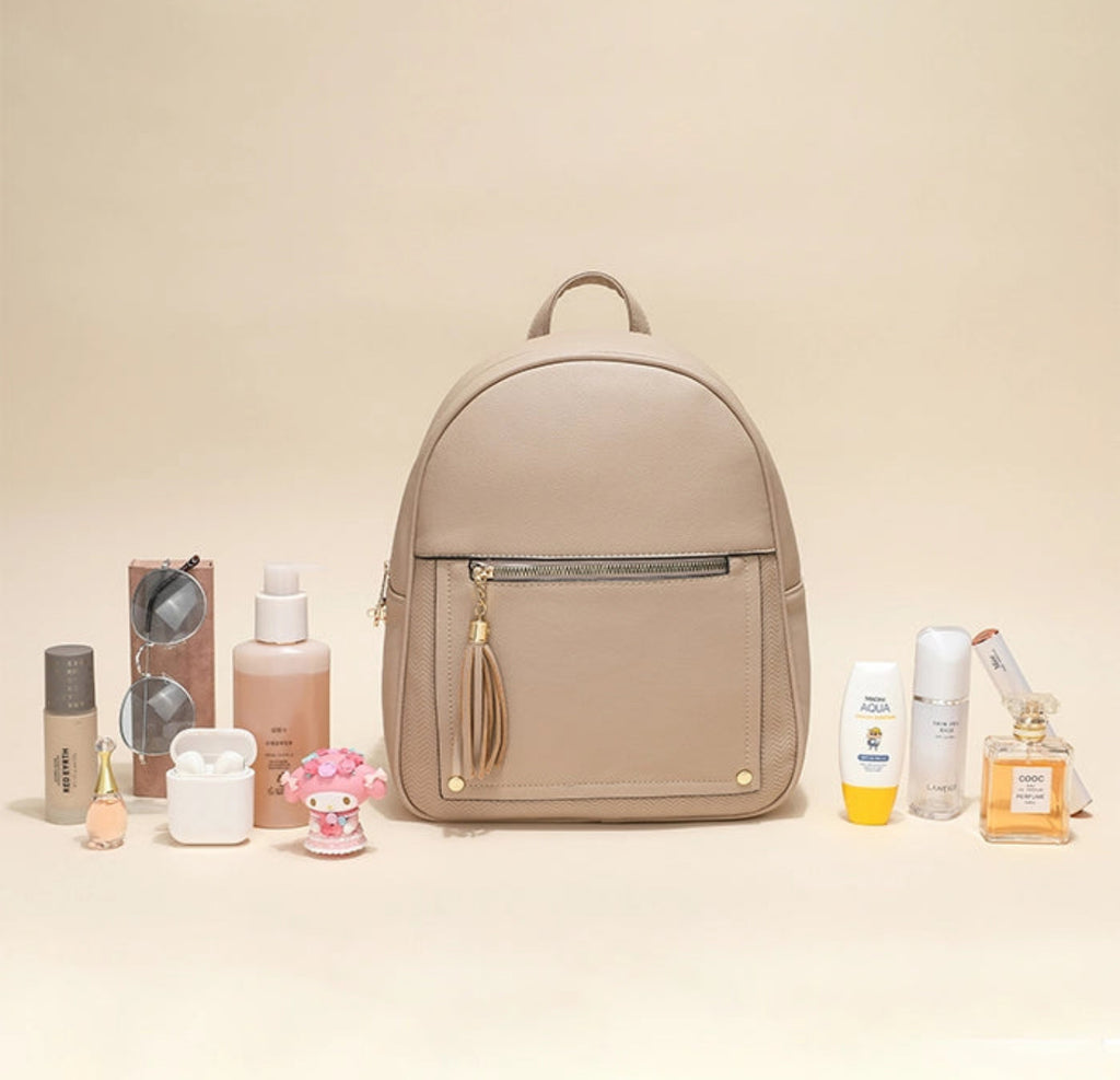 A beige faux-leather backpack with a front zip pocket and a tassel zipper pull, styled in a flat-lay arrangement with various beauty and perfume products on a warm beige background.